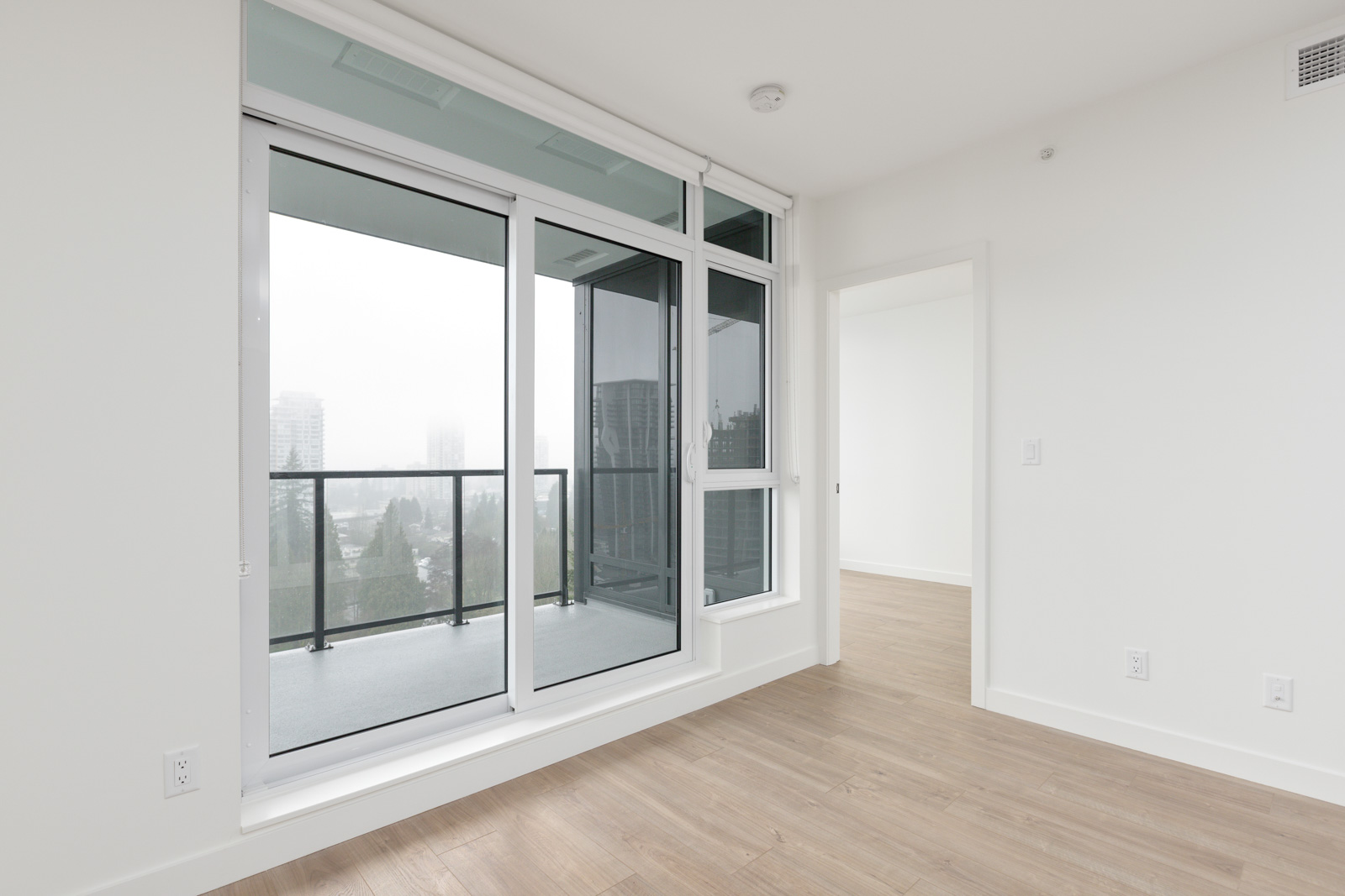 A modern empty room with light wood flooring, large glass sliding doors leading to a balcony, and an adjacent doorway to another room.