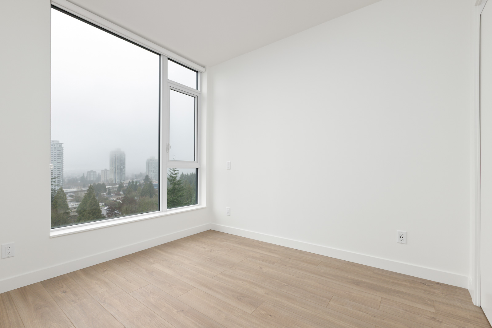 Empty room with light wood flooring, white walls, and a large window showing a cityscape with tall buildings and trees on a cloudy day.