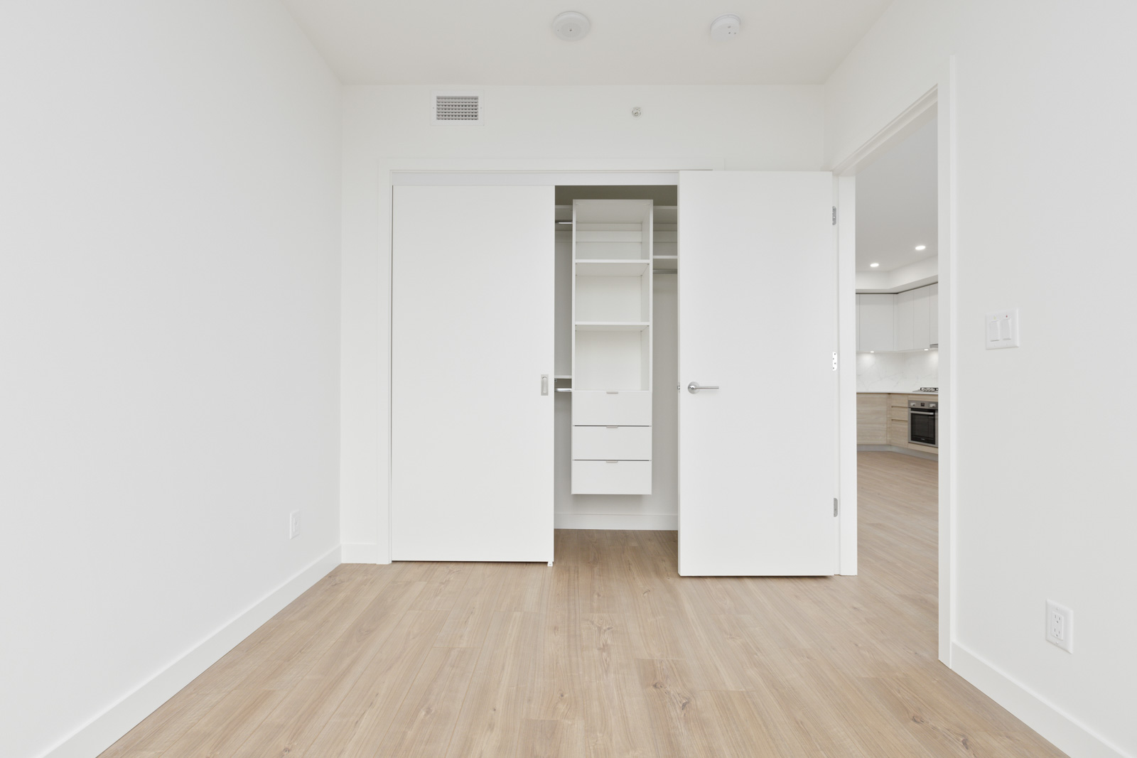 Empty room with light wood floors, white walls, and an open closet with shelves and drawers; adjacent room with visible kitchen area in the background.