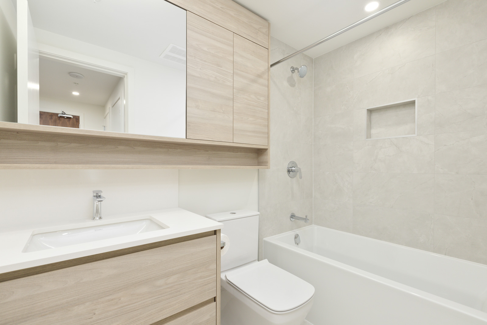 Modern bathroom with light wood cabinets, a white countertop and sink, a toilet, and a bathtub with a tiled shower surround and built-in shelf.