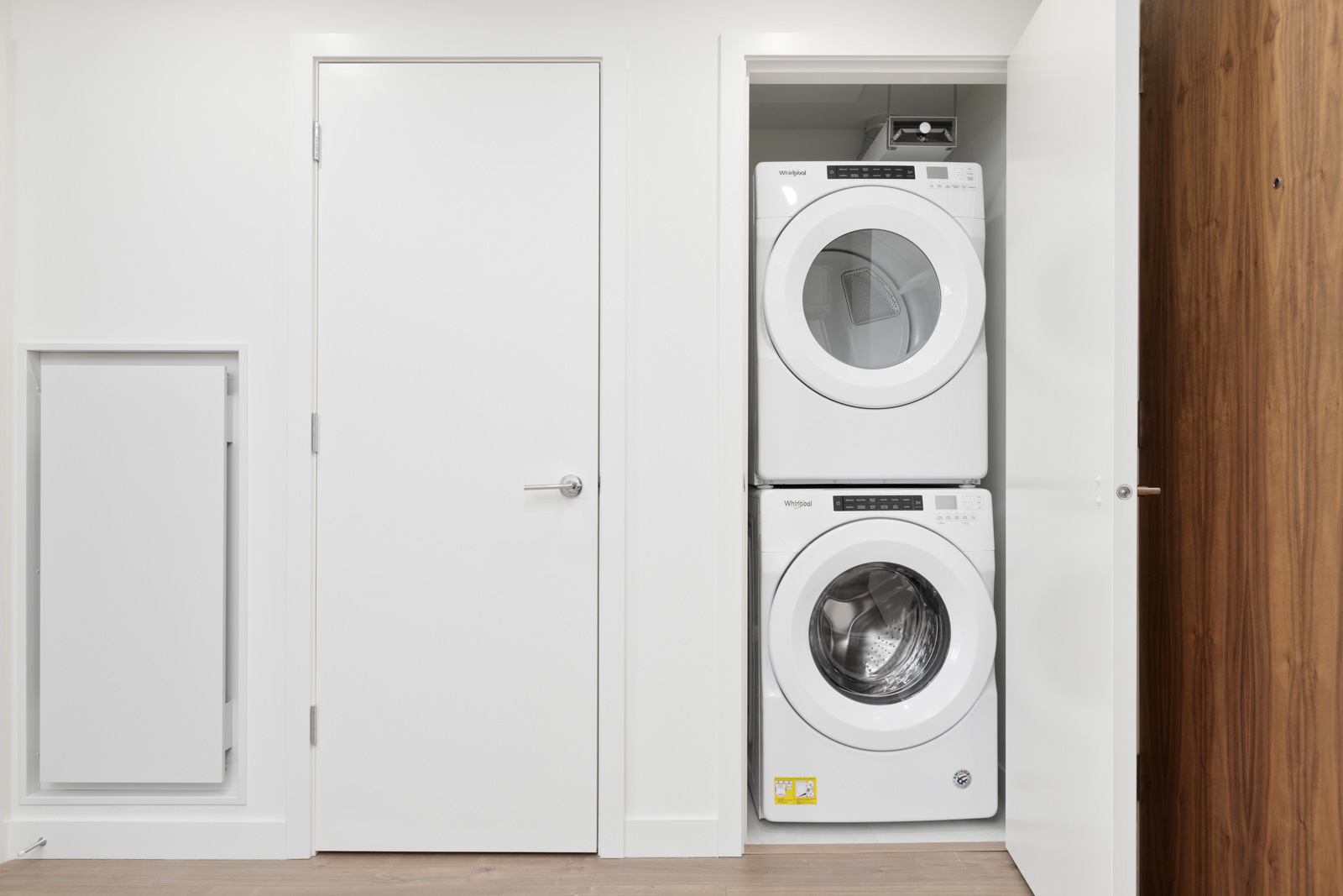 A stacked washer and dryer are installed in a closet with an open door in a modern laundry area with white walls and wood flooring.