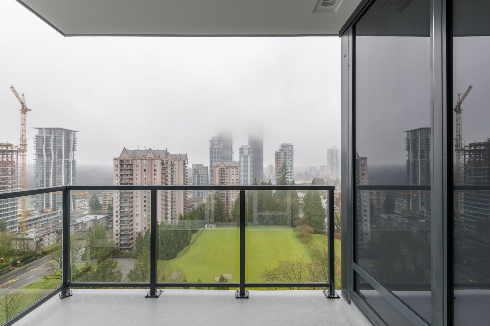 View from a high-rise balcony overlooking city buildings, construction cranes, and a grassy area on a foggy day.