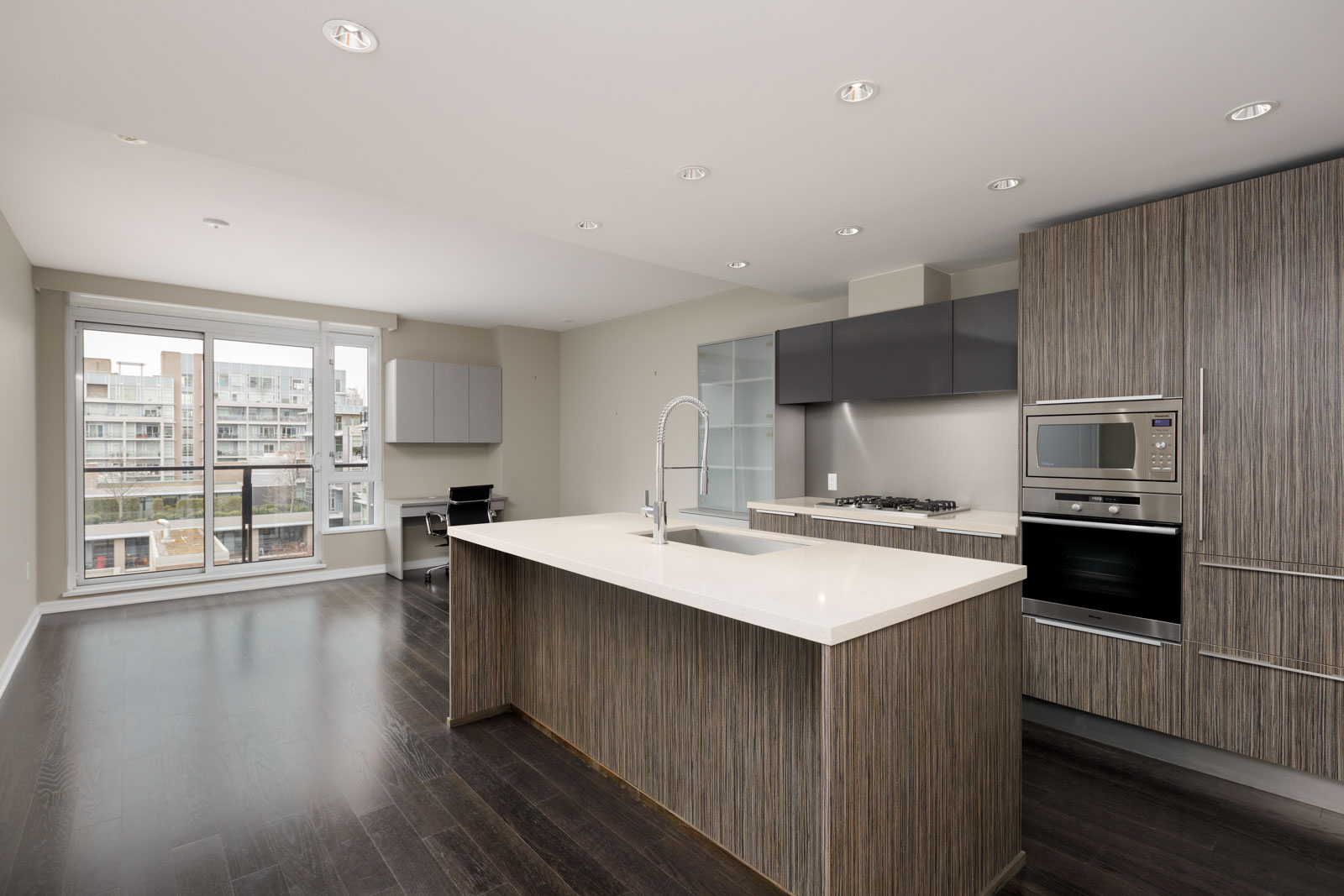Modern kitchen with dark wood floors, a large island with a sink, built-in stainless steel appliances, and floor-to-ceiling windows overlooking neighboring buildings.