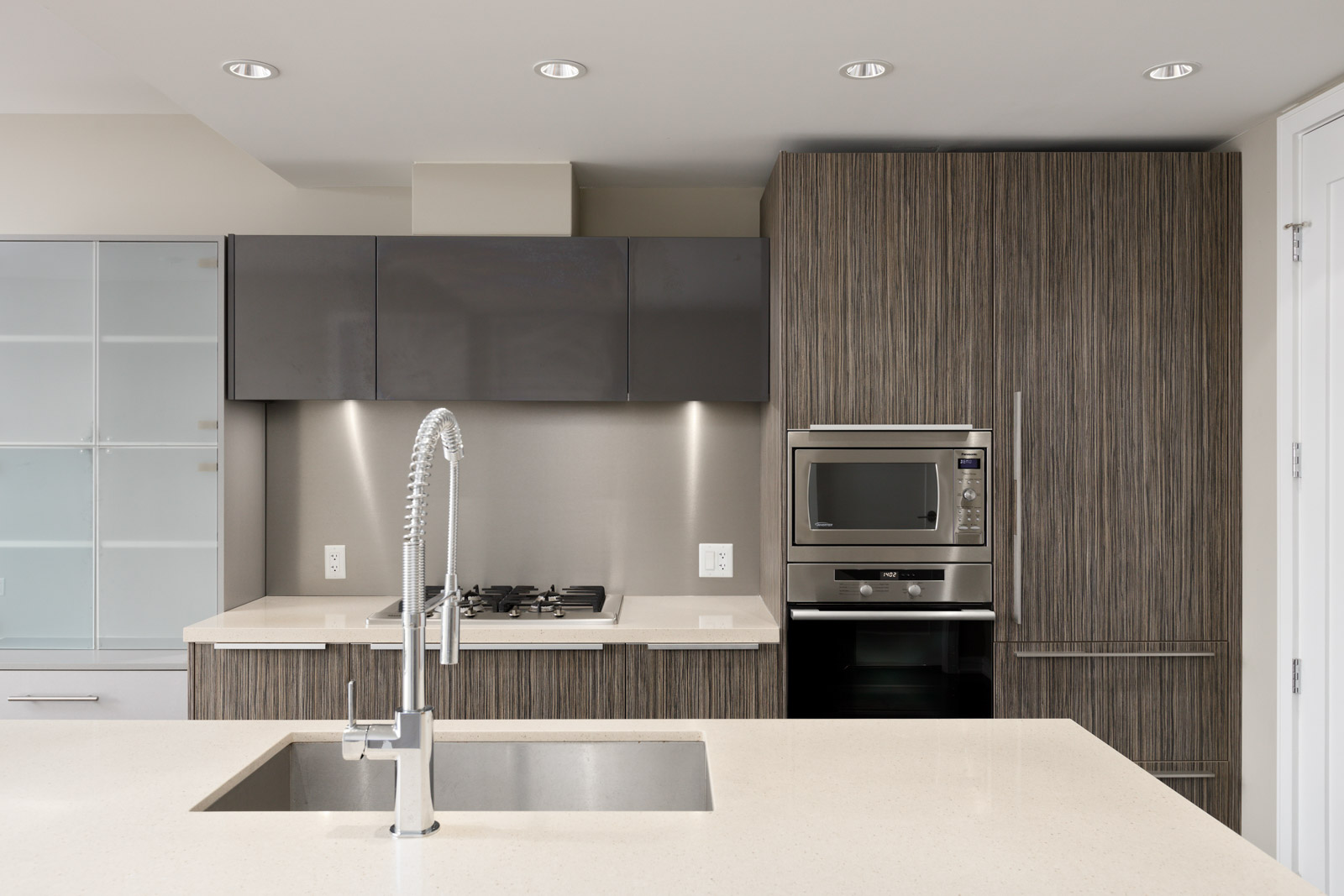 Modern kitchen with wood-textured cabinets, built-in oven, microwave, gas stove, under-cabinet lighting, and a central island featuring a sink and gooseneck faucet.
