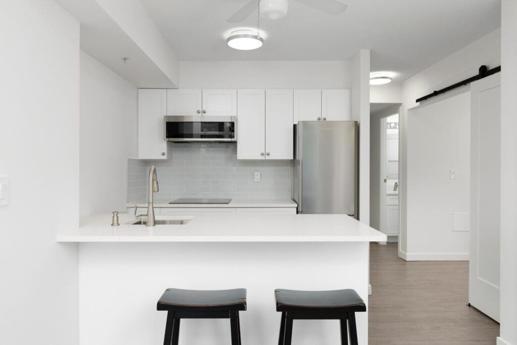 Modern white kitchen with a breakfast bar, two black stools, stainless steel appliances, subway tile backsplash, and wood flooring.