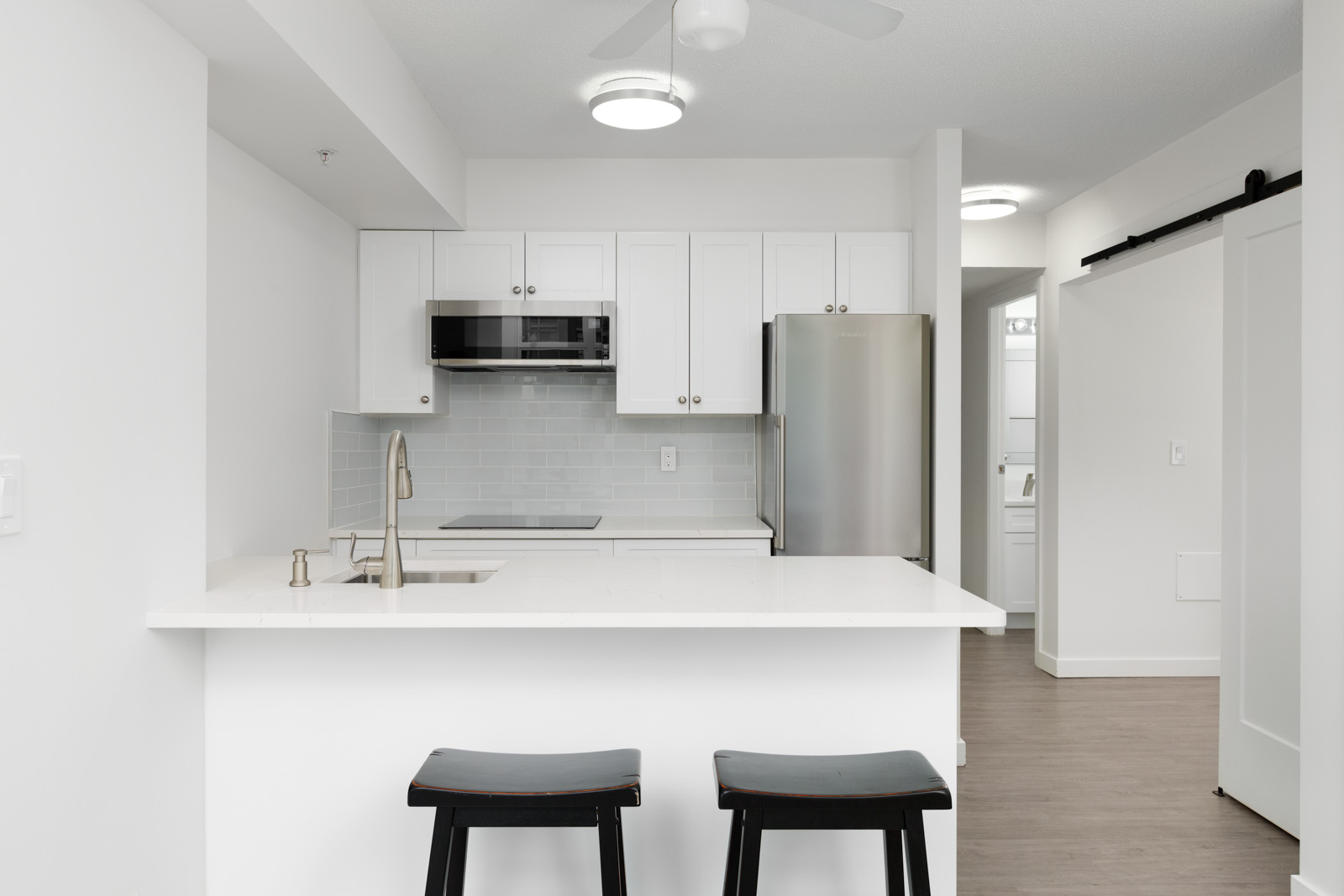 Modern white kitchen with a breakfast bar, two black stools, stainless steel appliances, subway tile backsplash, and wood flooring.