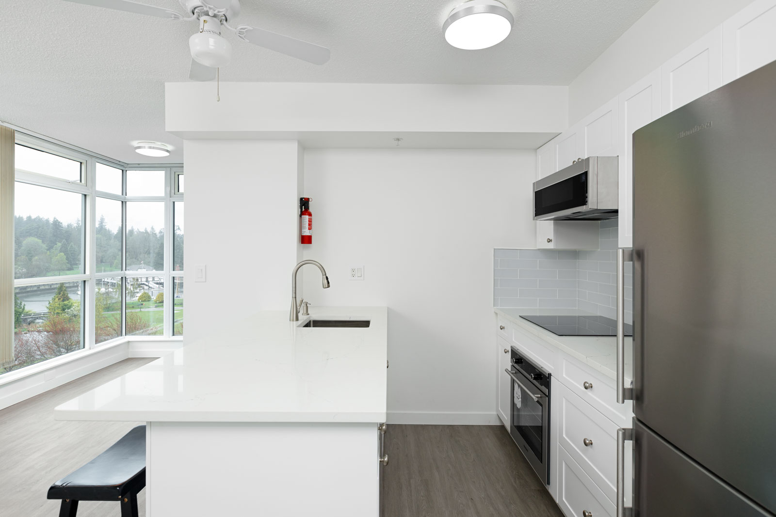 Modern kitchen with white cabinets, stainless steel appliances, and an island countertop. Large windows and natural light visible in the adjacent living area.