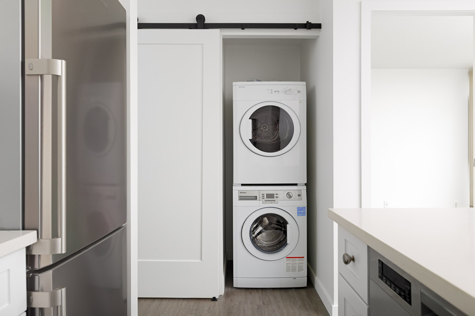 A compact laundry area with a stacked washer and dryer is partially hidden by a sliding white door next to a kitchen with stainless steel appliances.