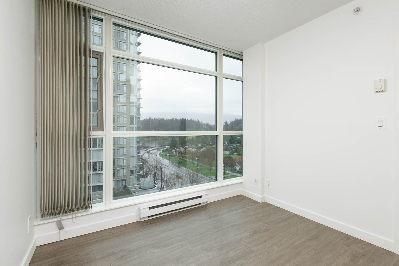 Empty room with light-colored walls, large floor-to-ceiling windows, vertical blinds, and a view of a high-rise building and greenery outside, with wood flooring.