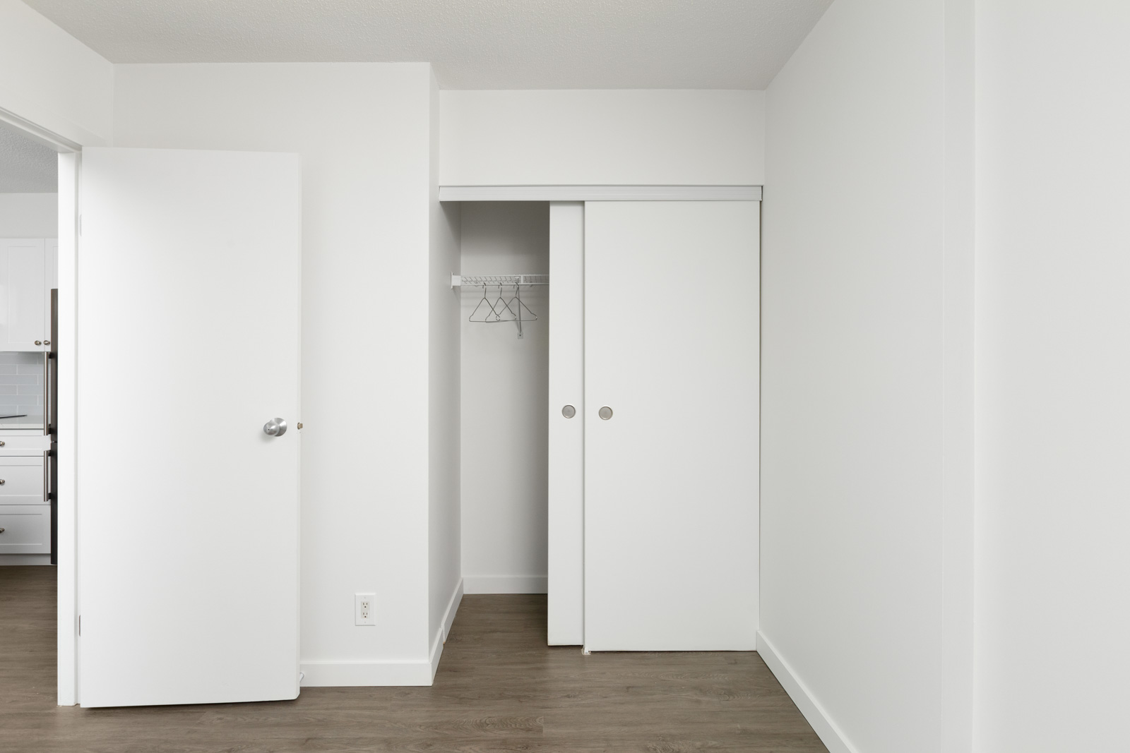 Empty white closet with sliding doors, a few hangers on a wire shelf, and an open door revealing part of a kitchen on the left. Hardwood floor throughout the space.