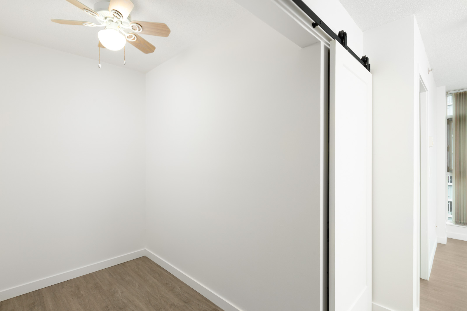 Small empty room with white walls, wood flooring, a ceiling fan with light, and a sliding barn door partially open to an adjoining space.