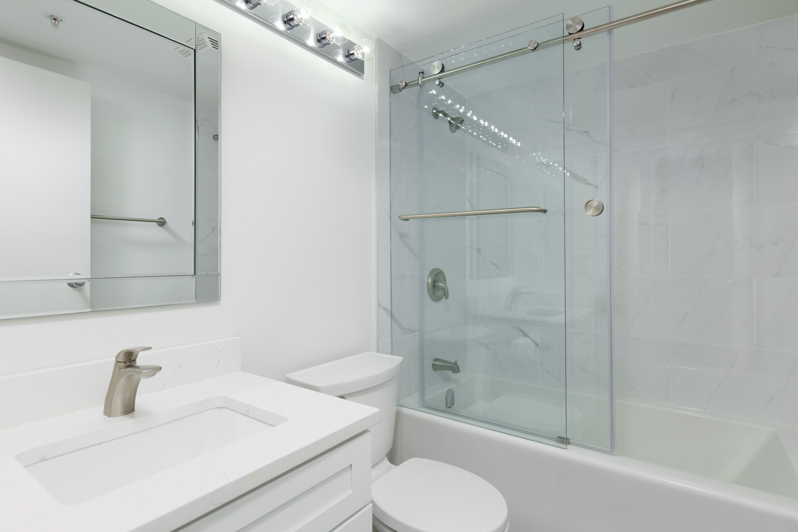 Modern bathroom with white walls, a sink with a faucet, a toilet, and a shower-tub combo with sliding glass doors and a metal grab bar.