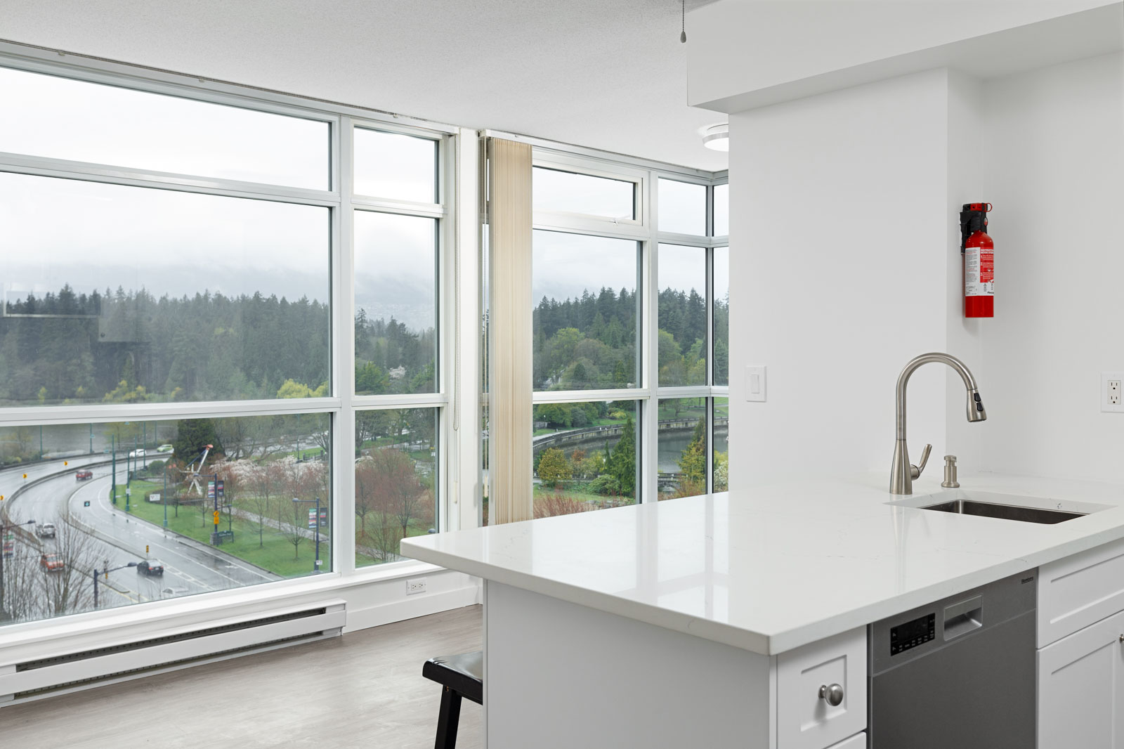 Modern apartment kitchen with white cabinets and an island, featuring large windows overlooking a forested area and a curving road.