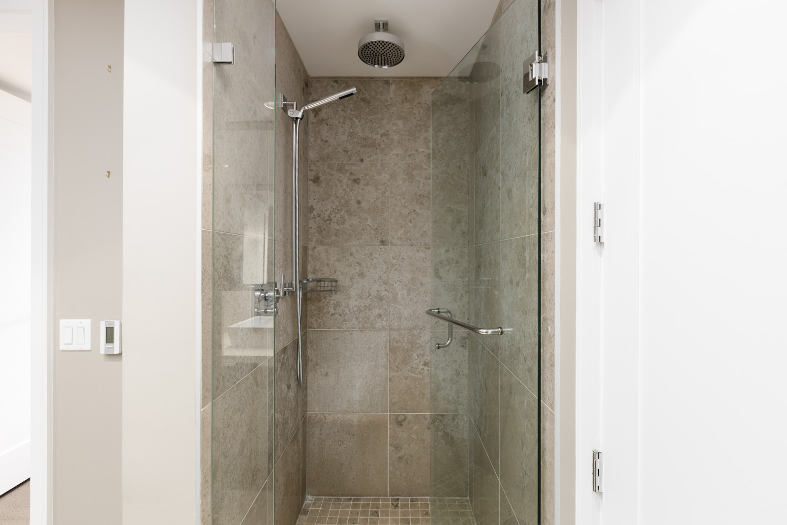 A modern walk-in shower with beige tiles, a glass door, a rain showerhead, and a metal grab bar on the right wall.