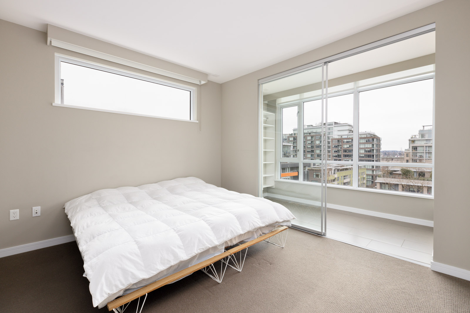 A minimal bedroom with a white bed, beige walls, carpeted floor, a high window, and sliding glass doors leading to a sunroom with city views.