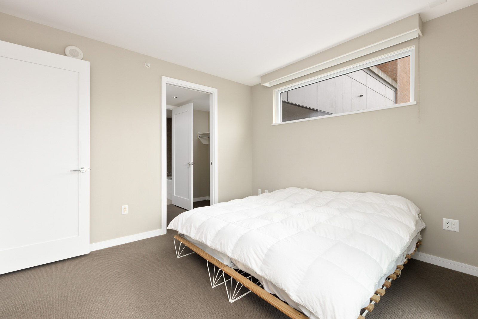 Minimalist bedroom with beige walls, a low bed with a white comforter, a window, and two white doors, one leading to a closet.