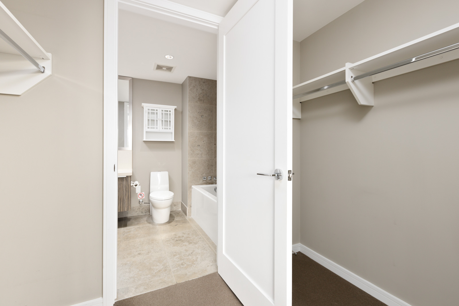 Open door reveals a modern bathroom with a toilet, bathtub, and white shelves, viewed from a carpeted walk-in closet with empty hanging rods.