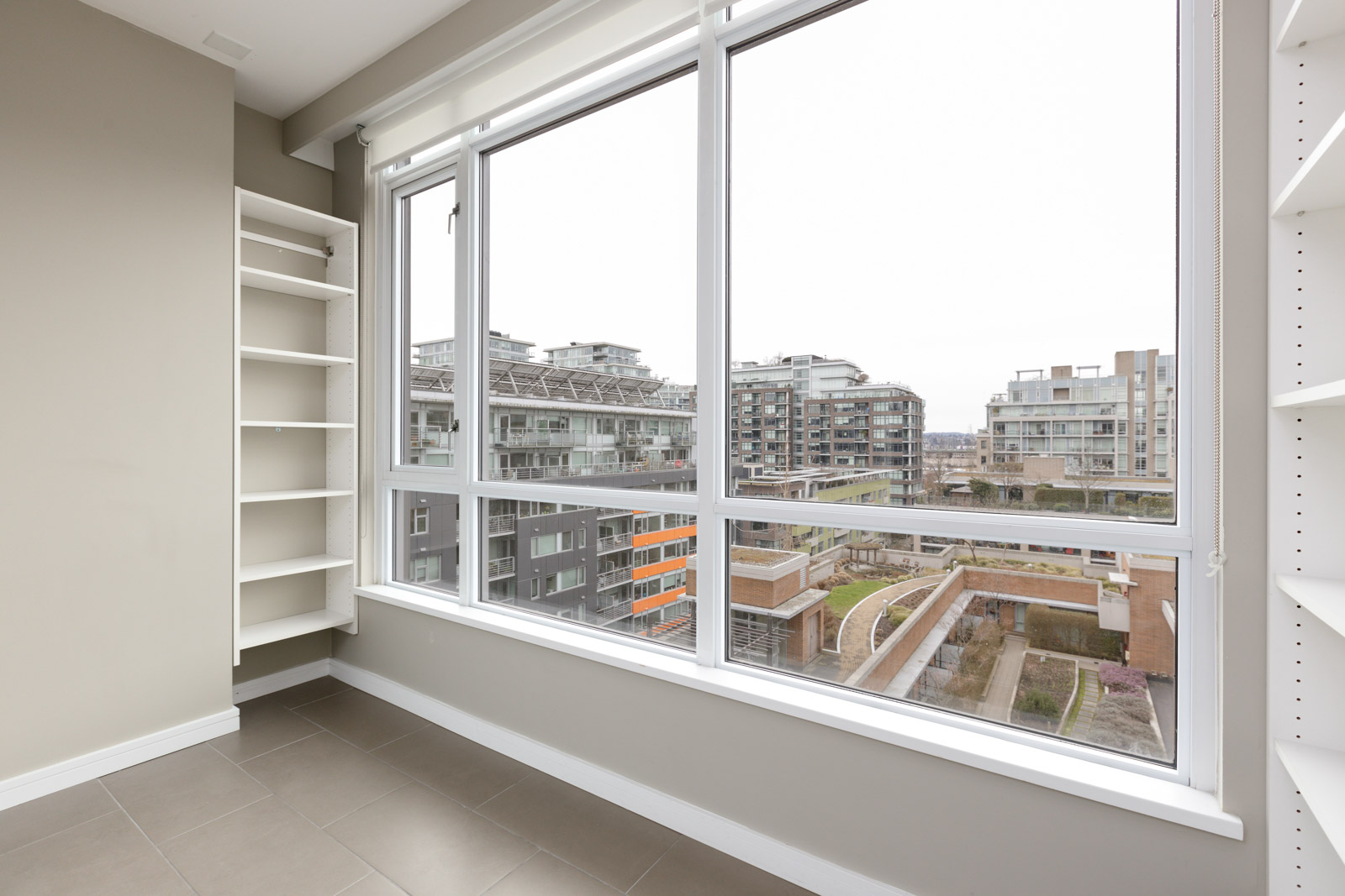 Empty, modern room with built-in shelves, large windows, and a view of nearby apartment buildings and rooftops.