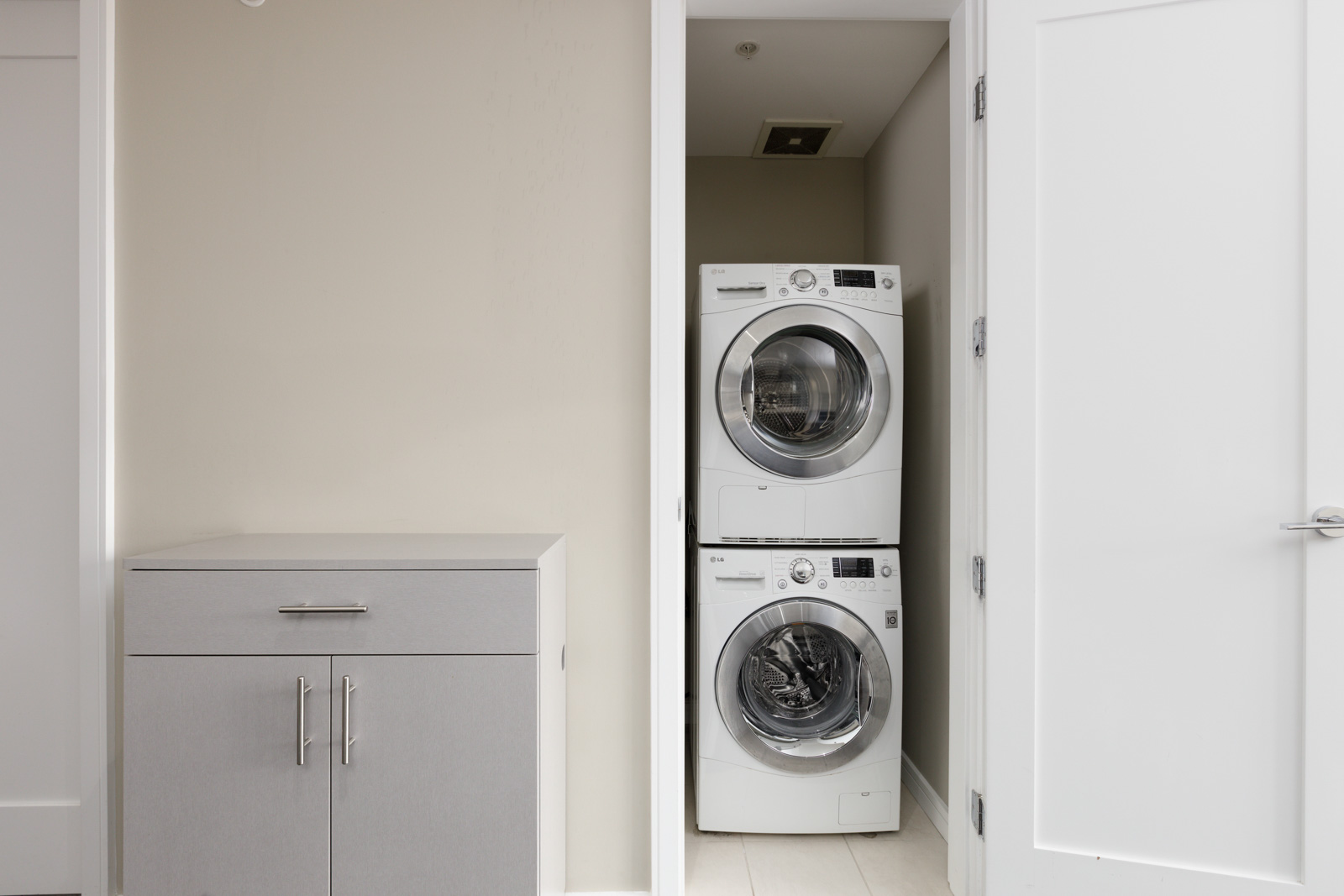 A stacked washer and dryer set is placed in a small laundry closet with white doors and beige walls next to a gray cabinet.