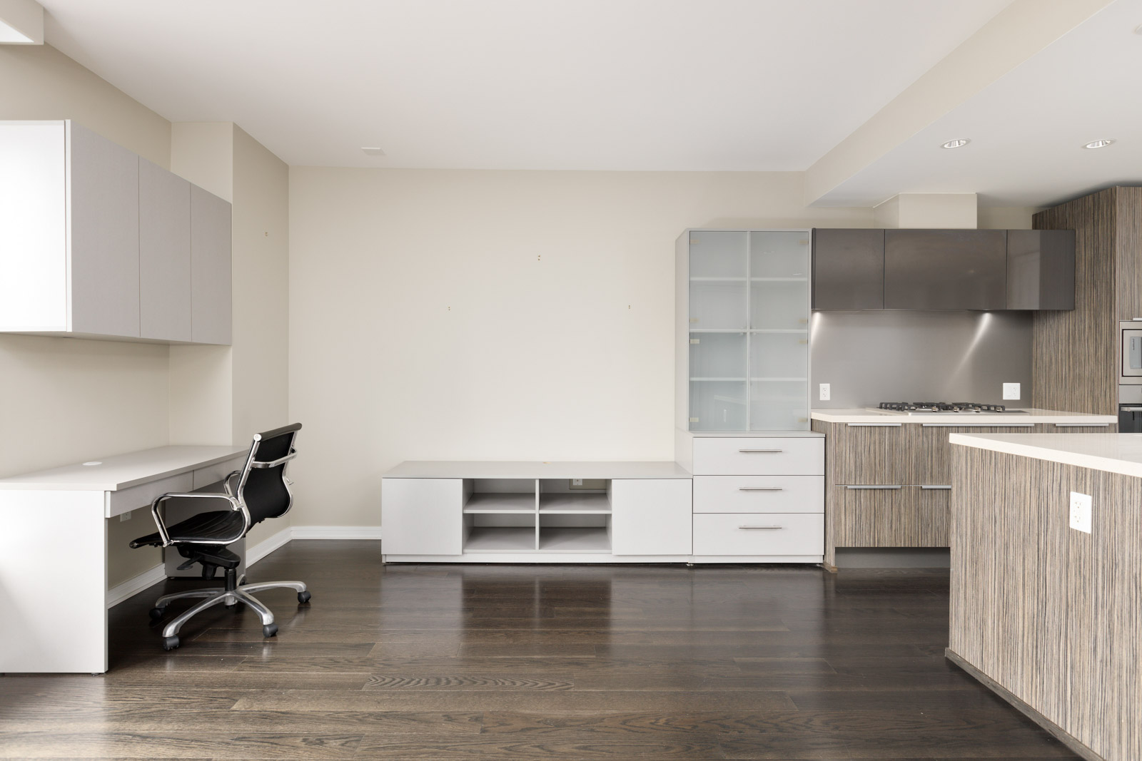 Modern kitchen and workspace with wood flooring, white cabinetry, a built-in desk with an office chair, and neutral wall colors.