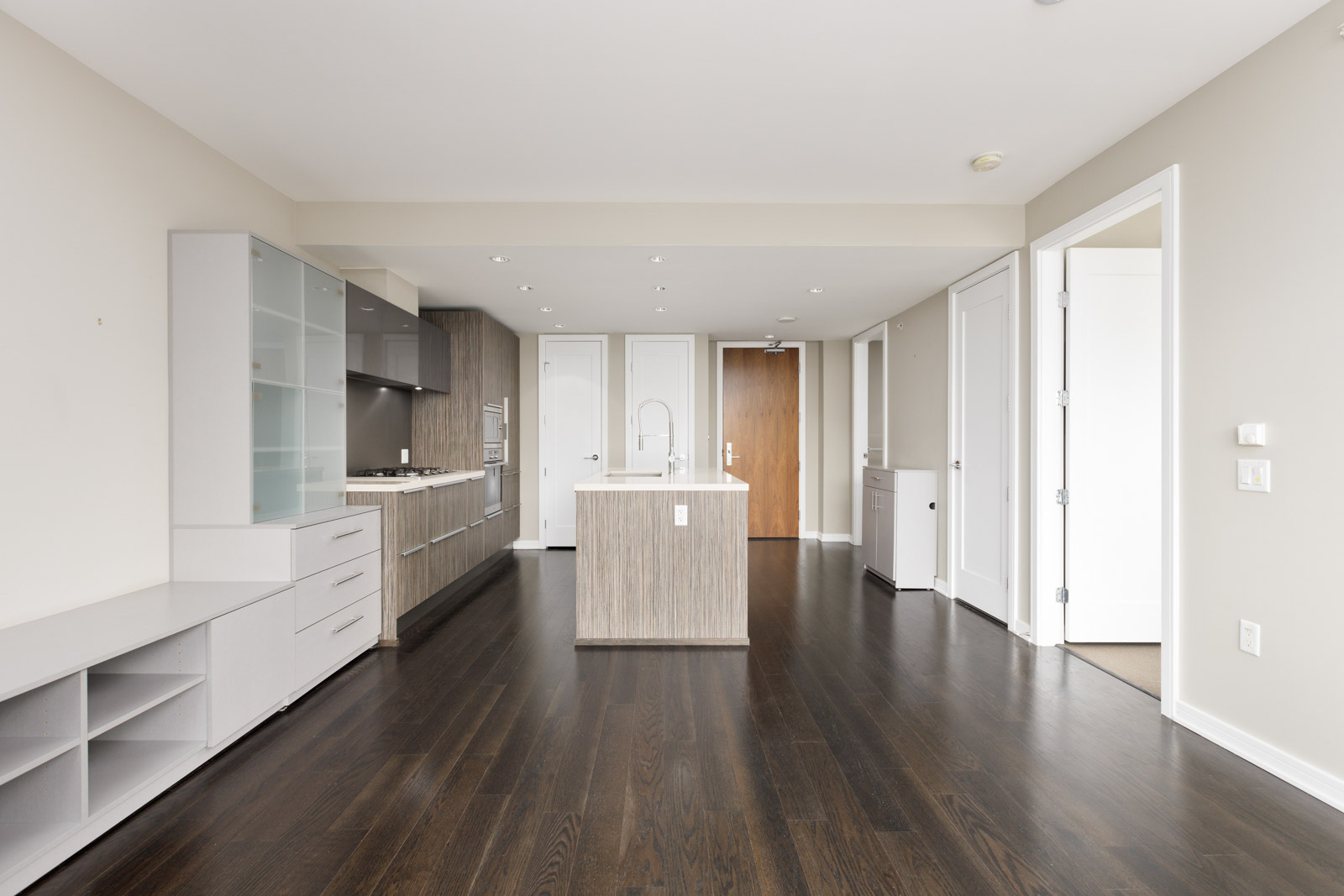 Modern apartment interior with dark hardwood floors, open kitchen with island, white walls, and several doors leading to other rooms.