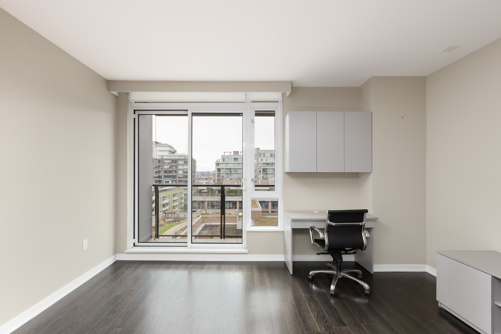 A modern, unfurnished room with dark wood floors, a glass door leading to a balcony, a built-in desk with cabinets, and a black office chair.