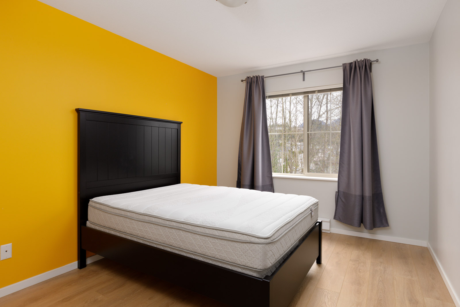 bedroom with a dark yellow accent wall. grey drapes for windows and bed in middle of room