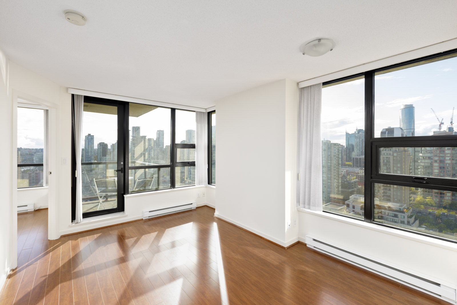 Bright, empty apartment room with large windows, hardwood floors, and a city skyline view. A balcony is visible through sliding glass doors on the left.