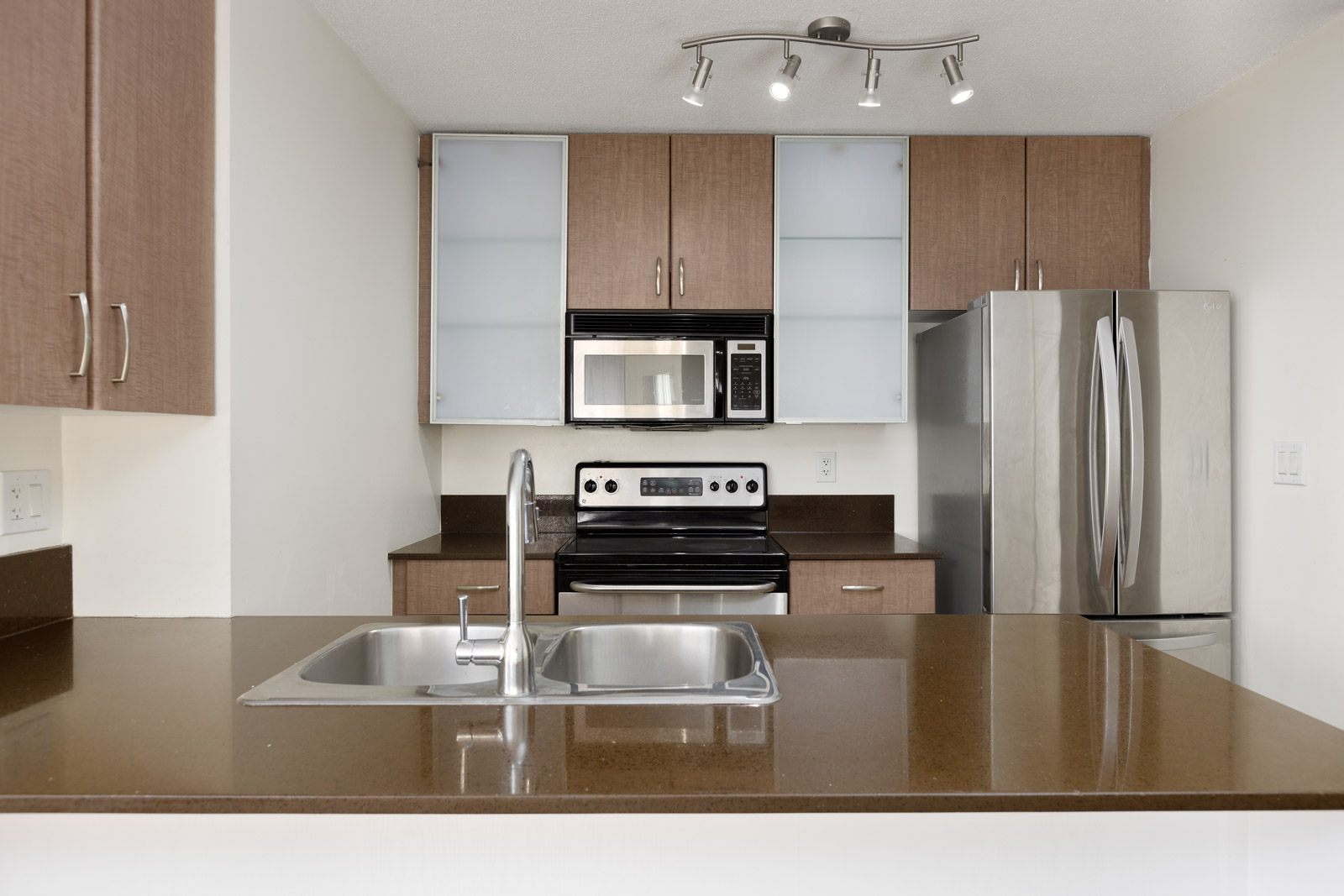 Modern kitchen with brown countertops, double sink, stainless steel refrigerator, oven with stove, microwave, and wood cabinets with frosted glass doors.