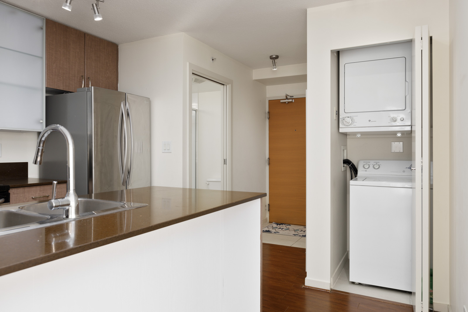 Modern apartment kitchen with brown countertops, stainless steel refrigerator, and a stacked washer and dryer unit in a closet near the entrance door.