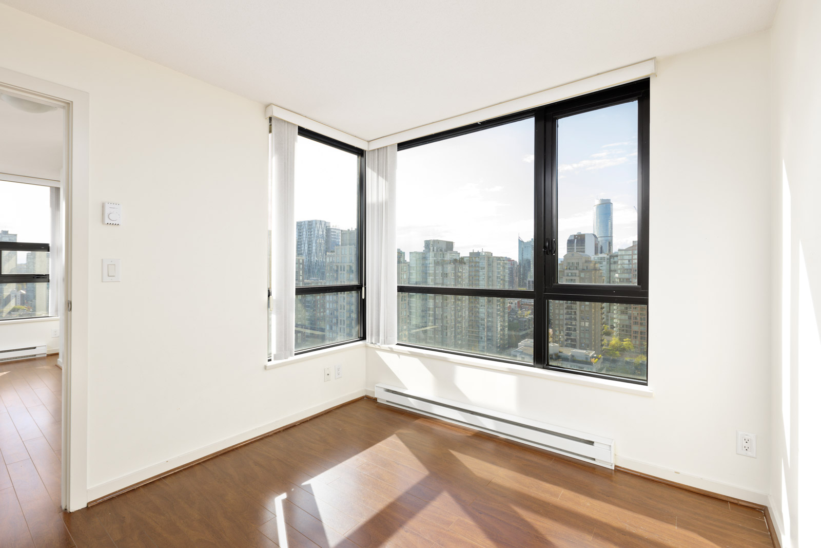 Unfurnished corner room with large windows, white walls, and wood flooring, overlooking a cityscape with tall buildings and clear sky.
