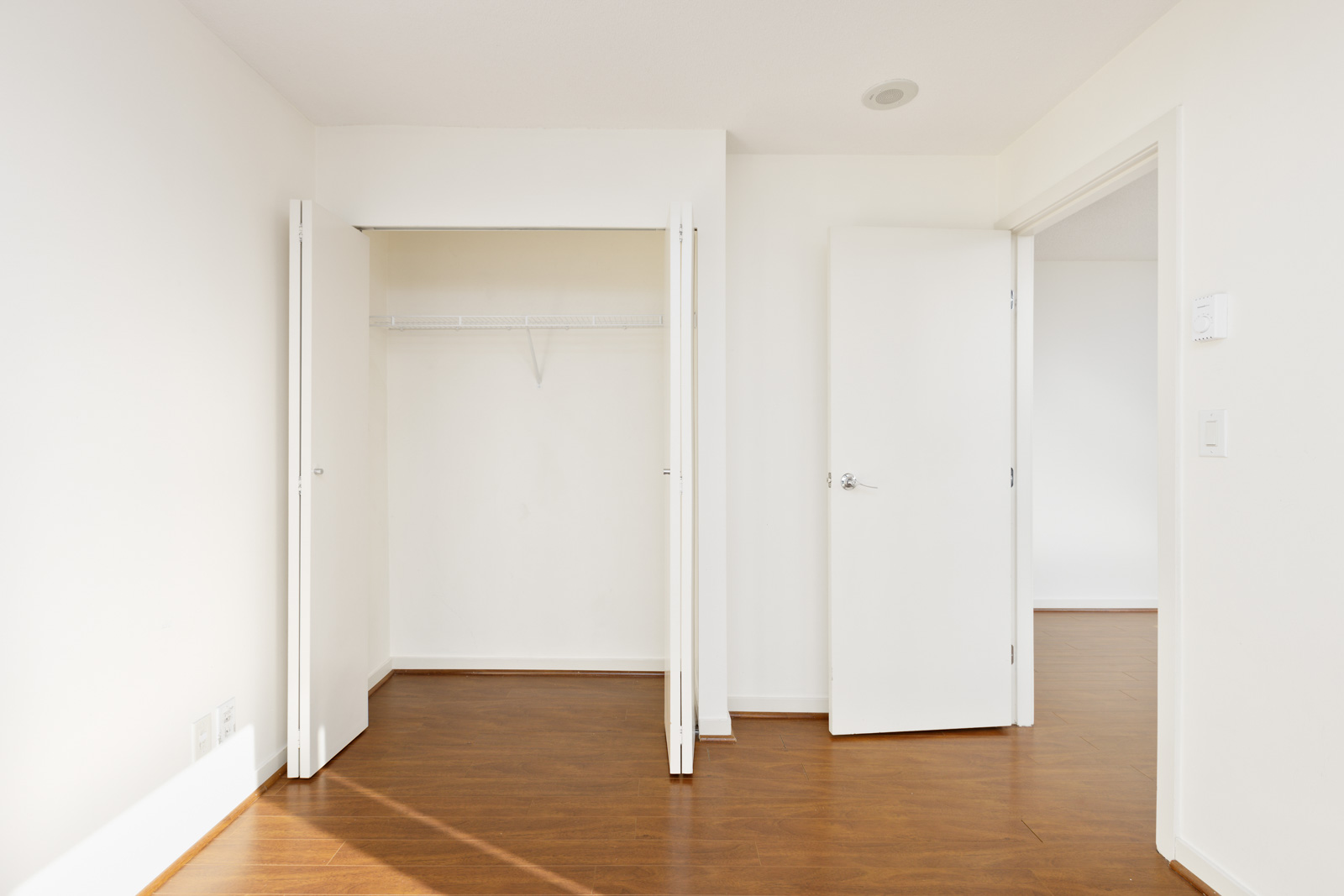 Empty room with white walls, open closet door showing a shelf and rod, brown wood flooring, and an open door leading to another room.