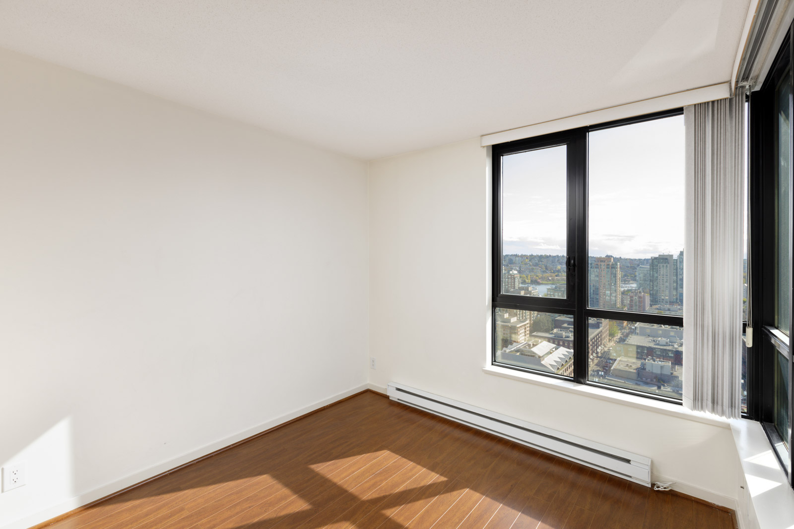 Empty, unfurnished room with light-colored walls, wood flooring, and large windows offering a city view with buildings and a partly cloudy sky.