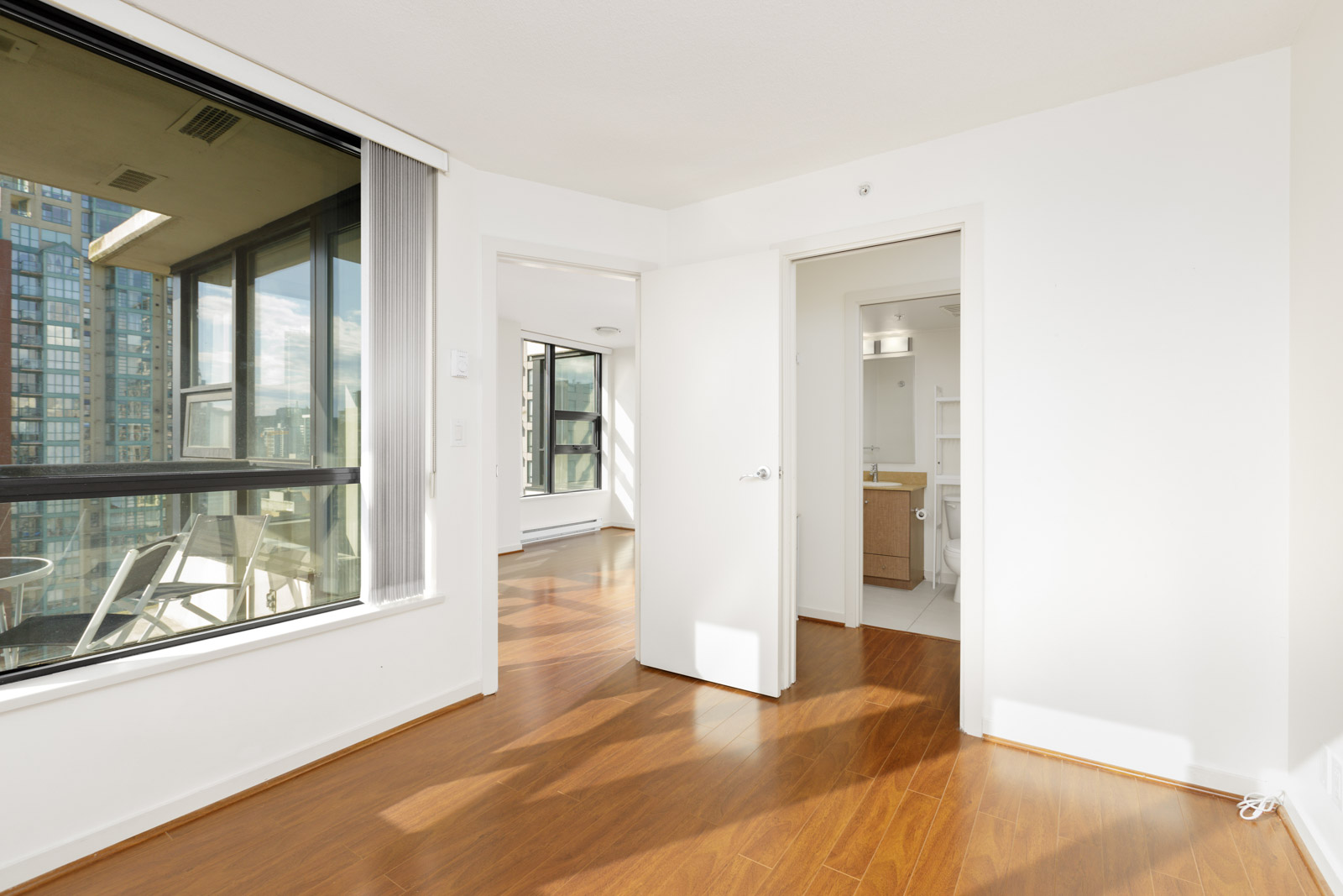 Bright, unfurnished room with hardwood floors, large window, sliding glass door to a balcony, and open doors leading to a bathroom and another room.