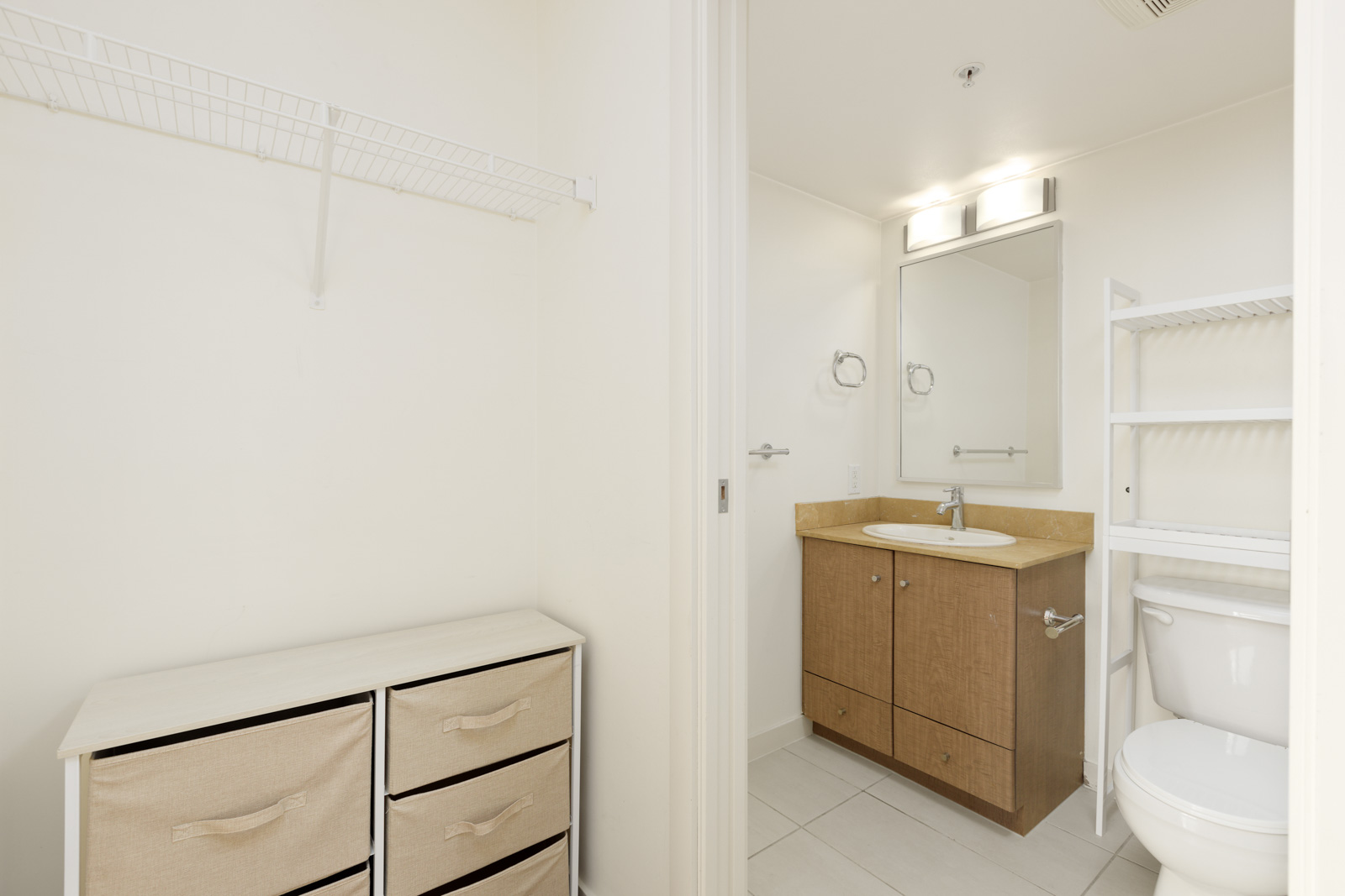 A bathroom with a wooden vanity, mirror, toilet, white shelving, tiled floor, and an adjacent area with a wire shelf and fabric drawer unit.