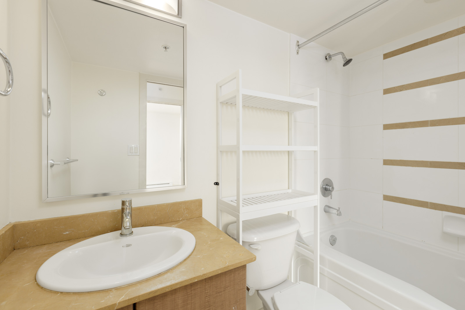 A bathroom with a sink, vanity, wall mirror, toilet, white shelving unit, and a bathtub with a showerhead and tiled walls.