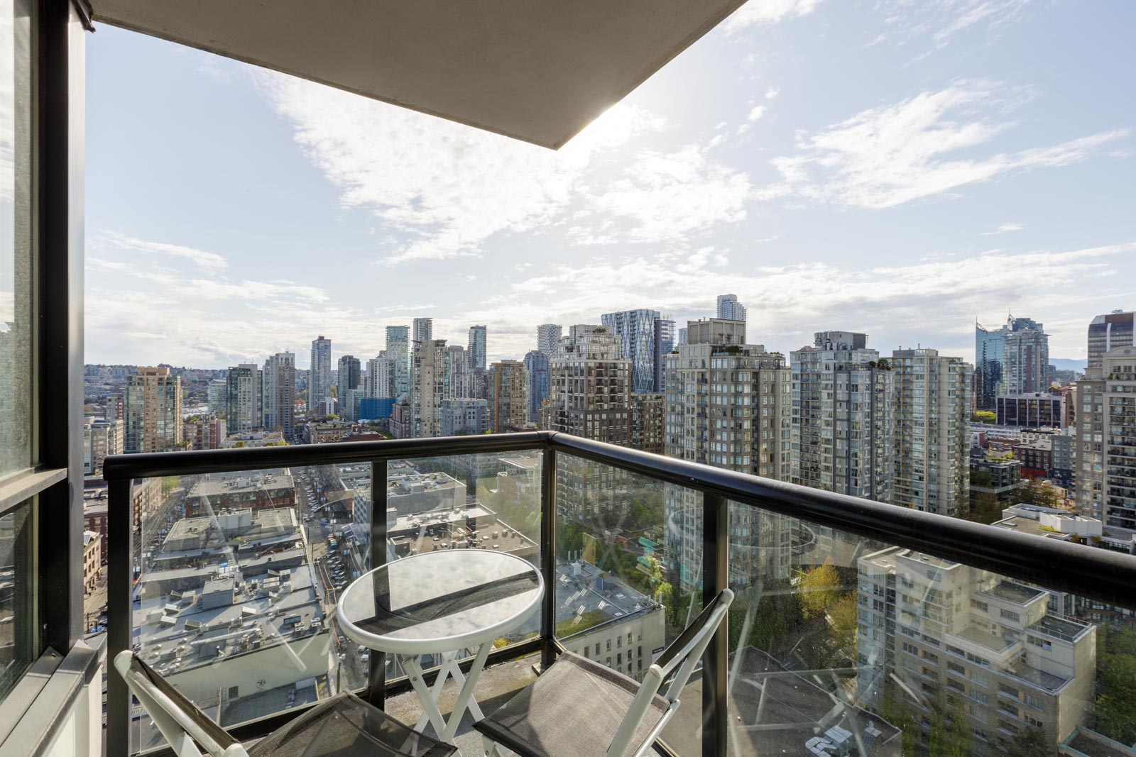 Balcony with glass railing, a small round table, and two chairs overlooking a cityscape of high-rise buildings under a partly cloudy sky.