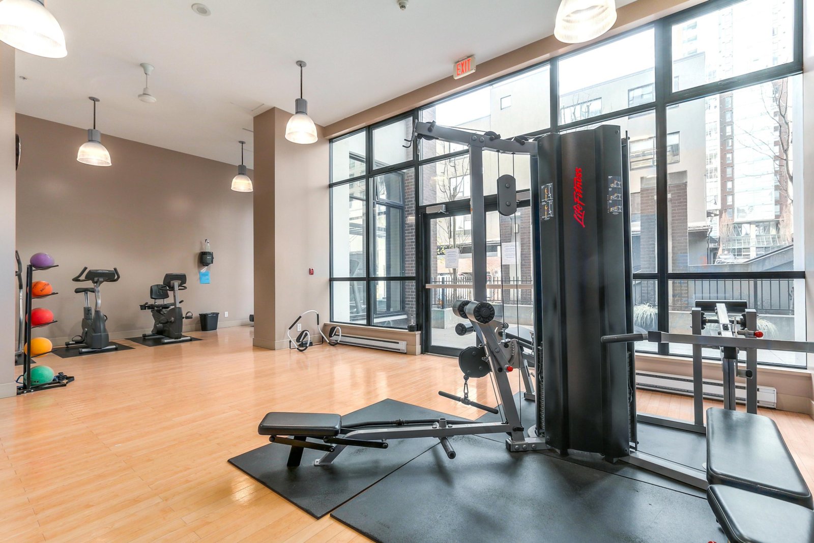 Modern gym with large windows, various exercise machines, free weights, and yoga balls on a wooden floor.