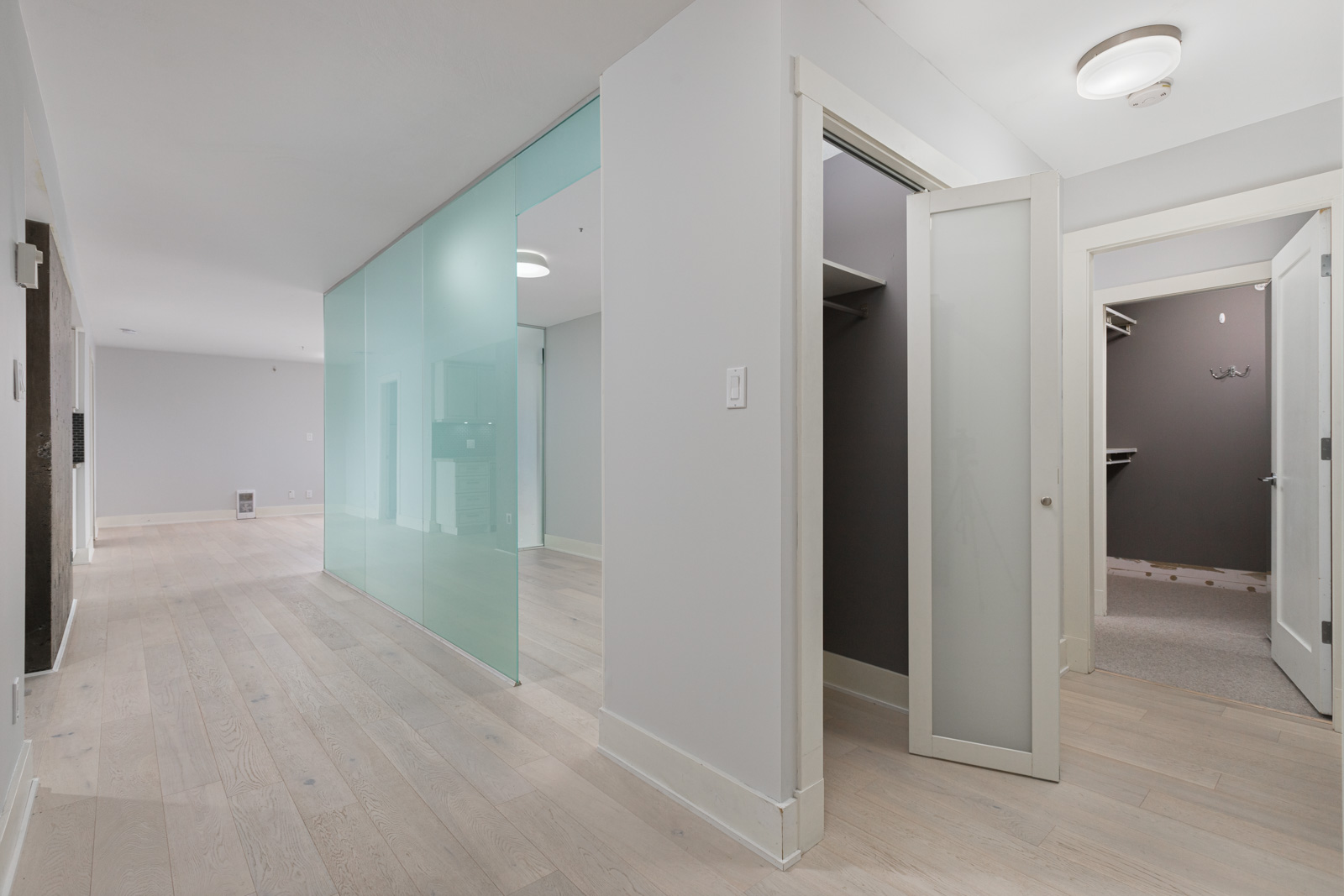 A modern hallway with light wood flooring, a frosted glass wall, and open doors leading to walk-in closets with built-in shelving.