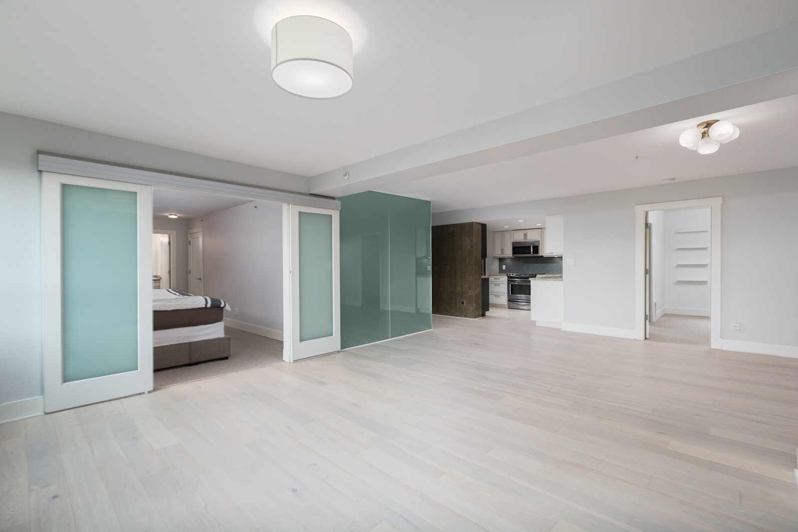 Spacious modern apartment with light wood floors, a bedroom separated by sliding glass doors, open kitchen, and built-in shelves.