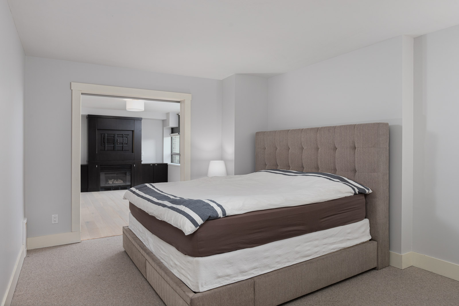 A bedroom with a neatly made bed, light gray walls, beige carpet, and a nightstand with a lamp; an open doorway leads to a room with a fireplace.