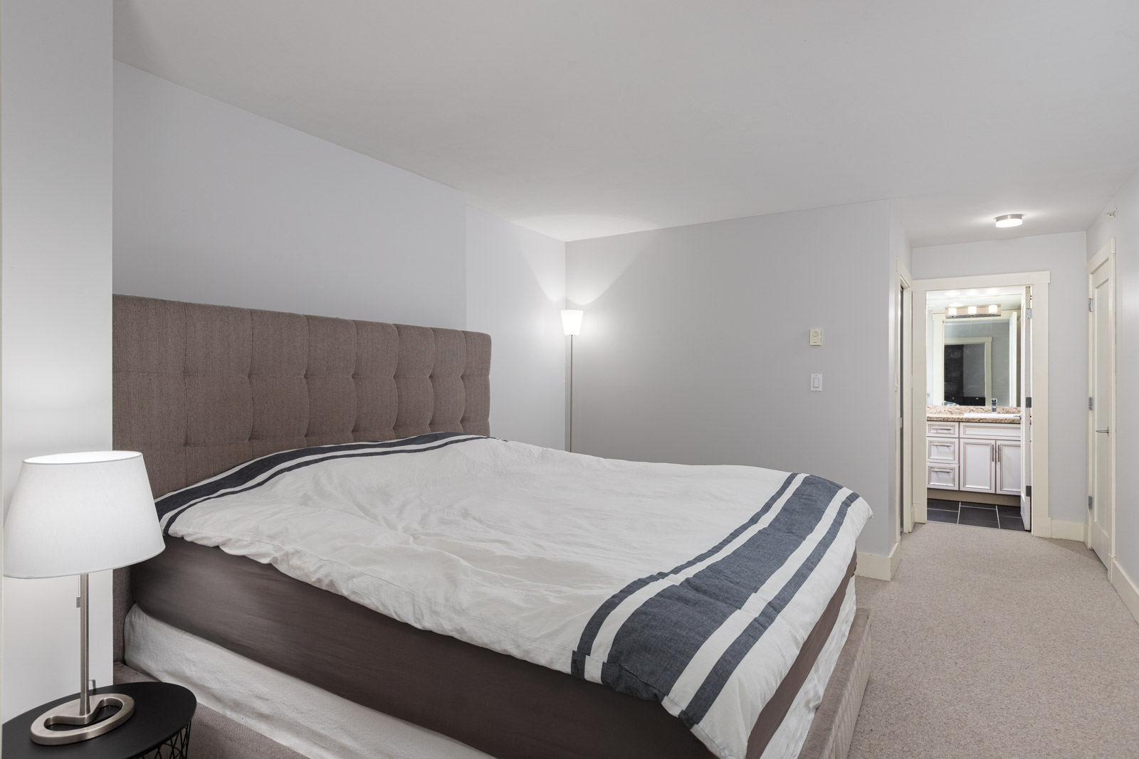 A modern bedroom with a large bed, two bedside lamps, light gray walls, and a view into a brightly lit bathroom with a vanity and mirror.