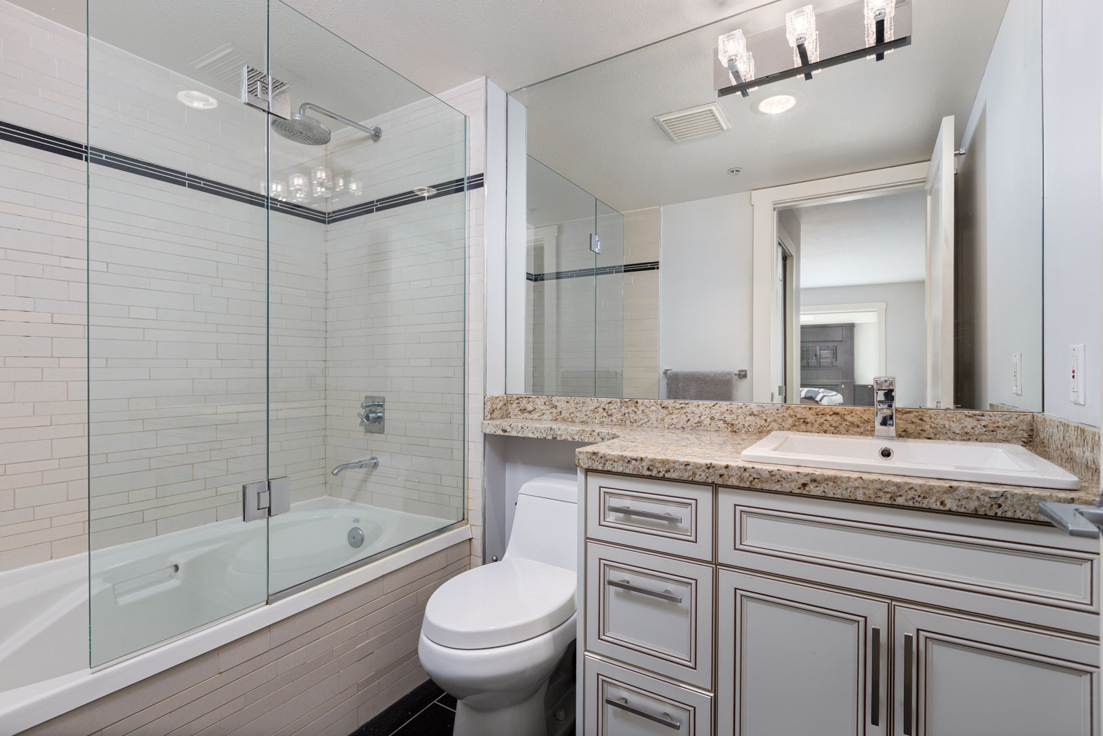 Modern bathroom with a glass-enclosed bathtub, toilet, granite countertop, white cabinets, sink, and a large mirrored wall.
