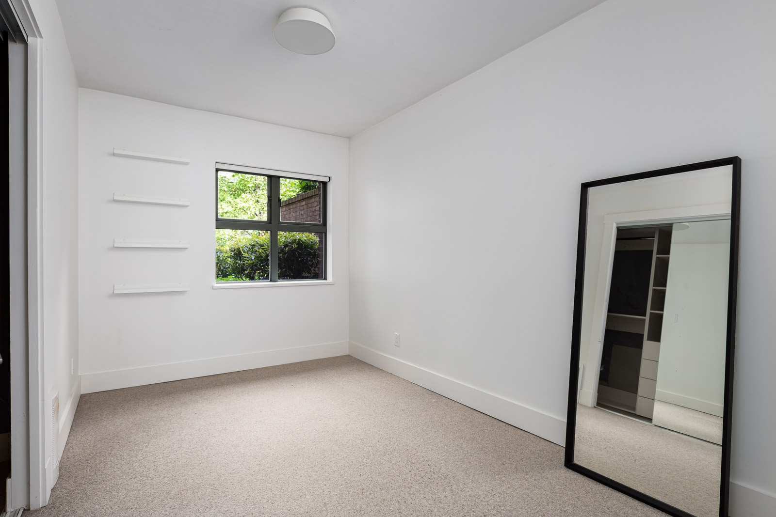 Empty room with white walls, beige carpet, a window, three wall shelves, and a large mirror leaning against the right wall reflecting part of a closet.