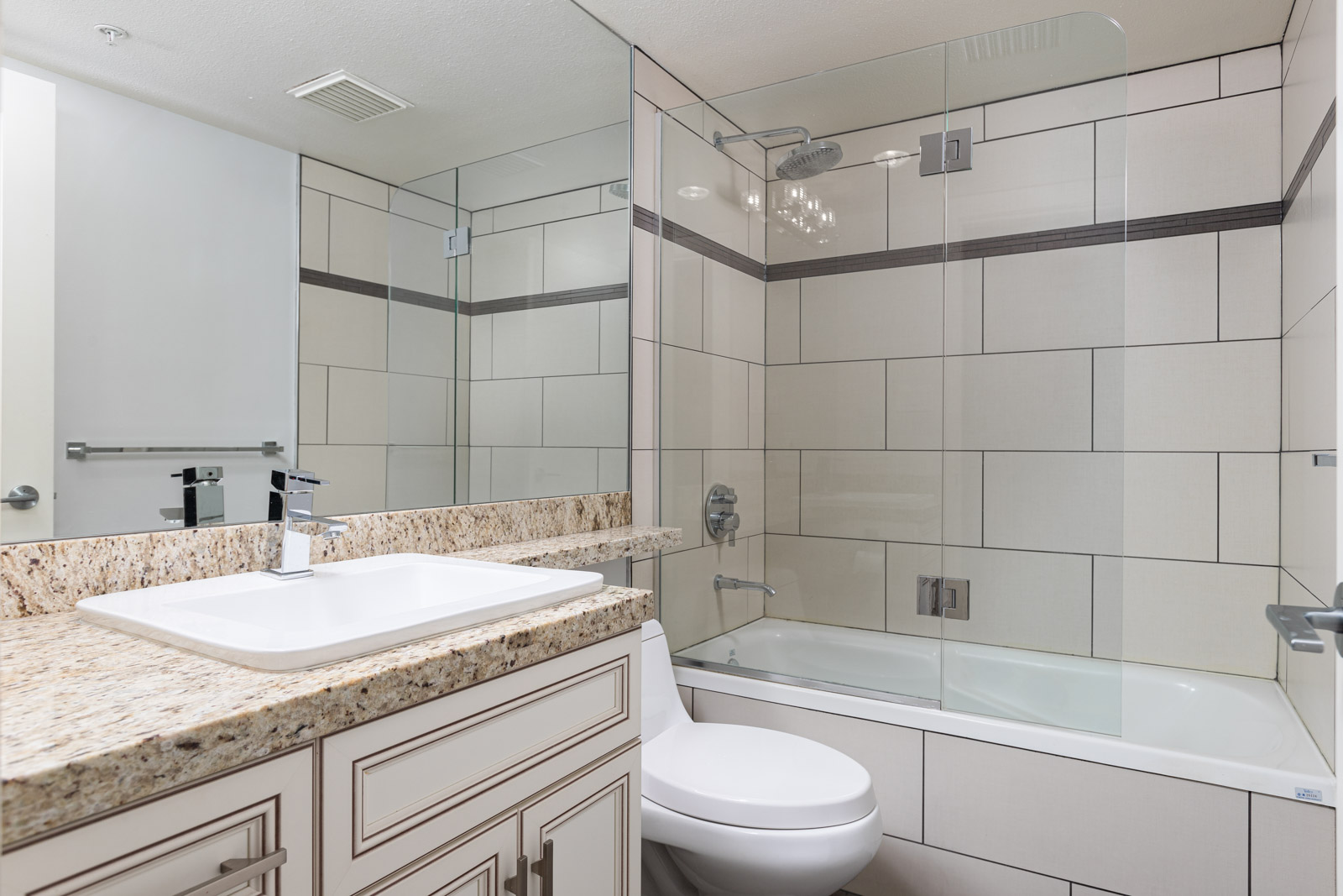 Modern bathroom with a granite countertop, white sink, toilet, and a glass-enclosed bathtub with tile walls and a large wall mirror.