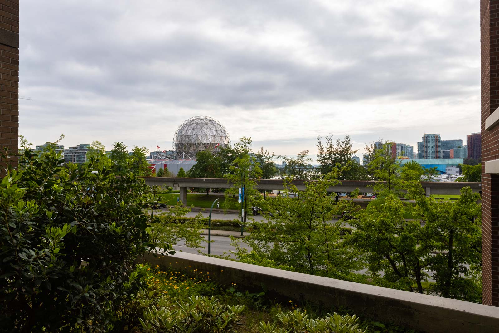 View of a cityscape with a geodesic dome structure in the background, surrounded by trees, buildings, and a road in the foreground under a cloudy sky.