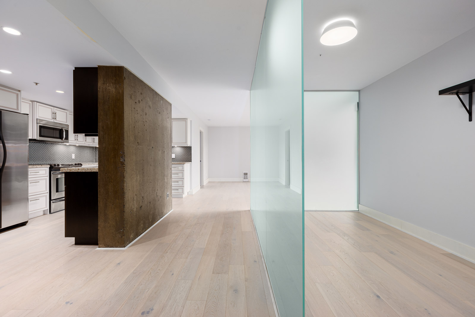 Modern kitchen with stainless steel appliances and white cabinets on the left, separated by a glass wall from an empty room with light wood flooring on the right.
