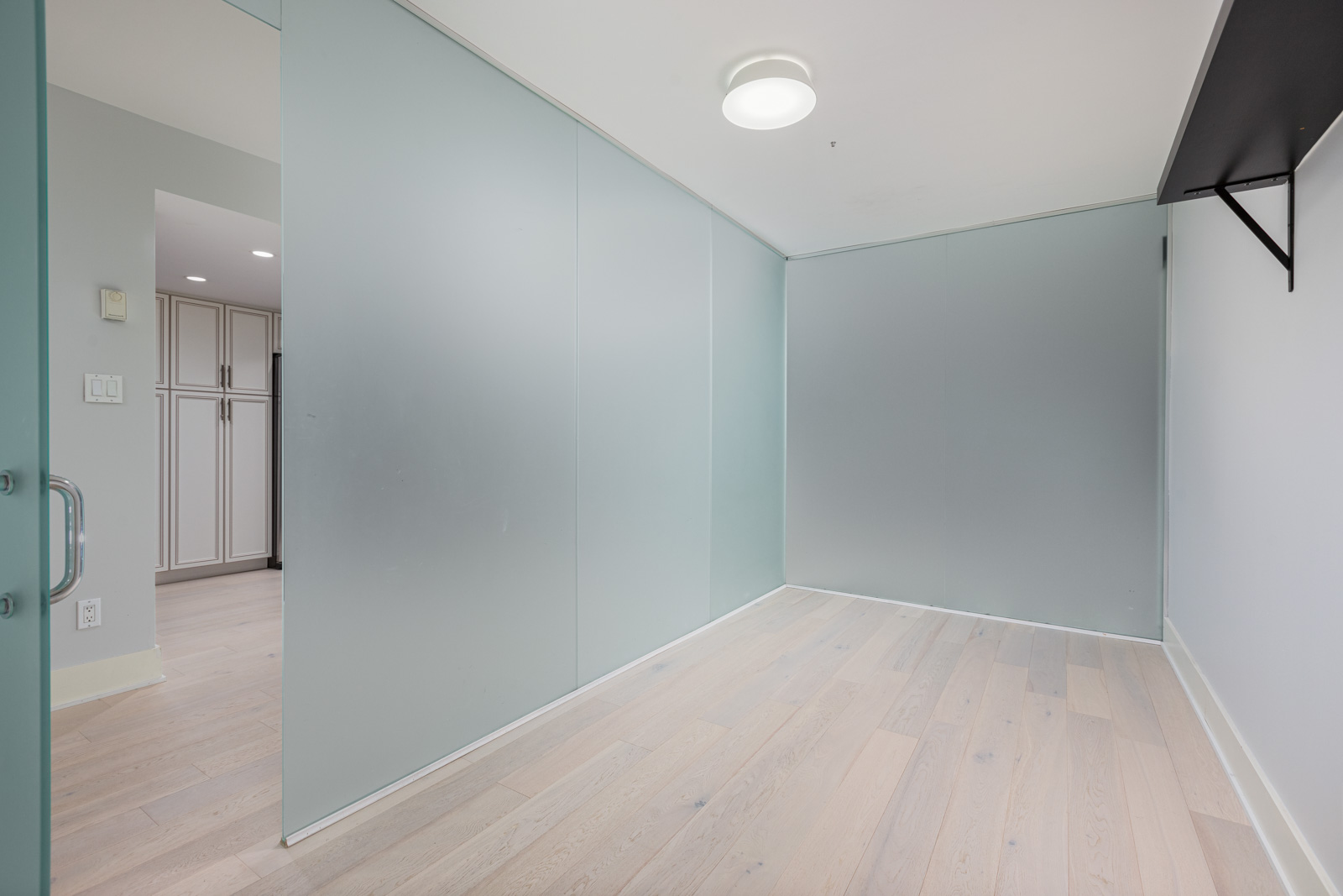 A small, empty room with frosted glass walls, light wood flooring, a black shelf on the right wall, and an open door leading to a kitchen area.