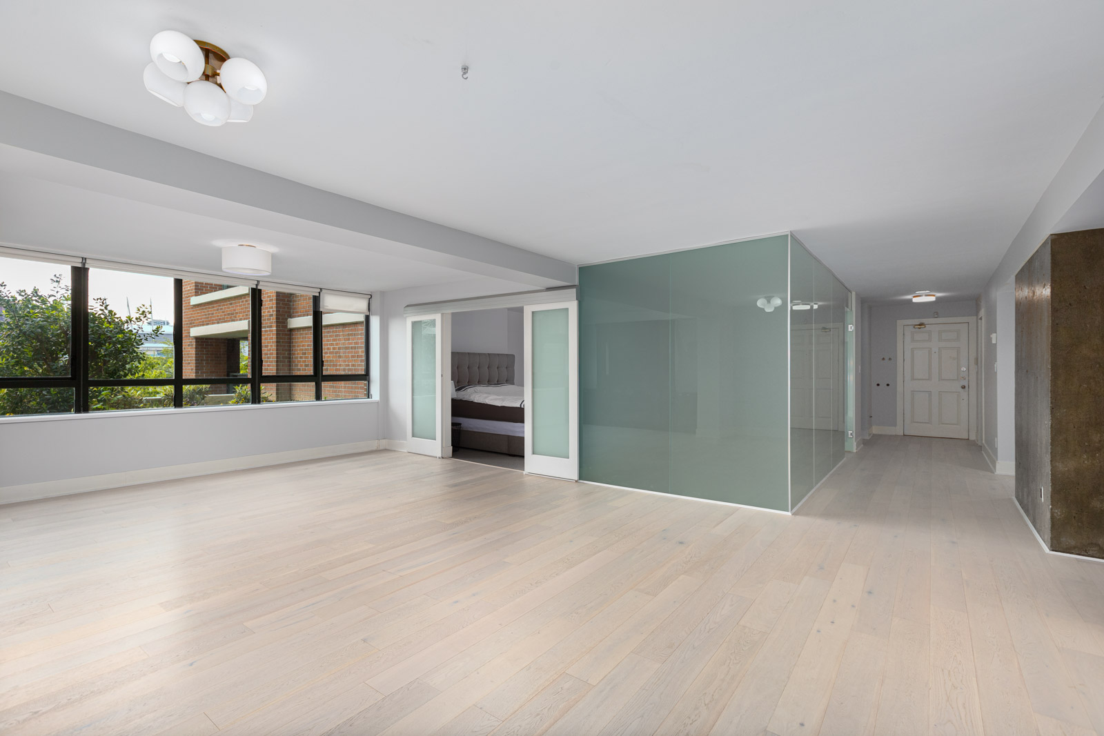 Spacious, modern apartment with light wood floors, large windows, glass-walled bedroom, and white ceiling lights.