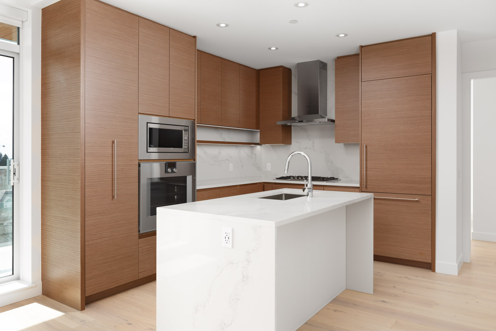 Modern kitchen with wood cabinets, built-in stainless steel appliances, white marble island with sink, and light wood flooring.